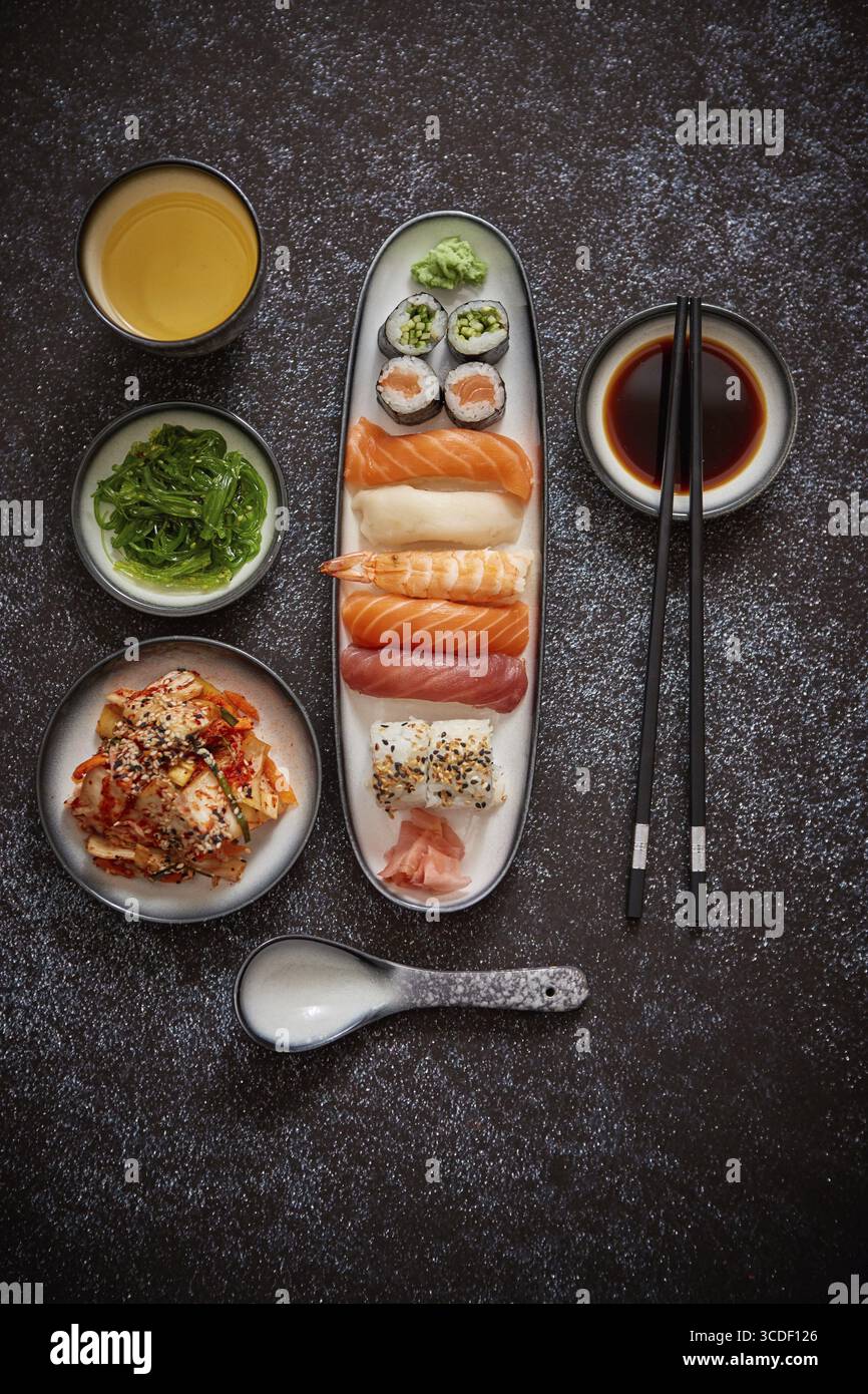 Assortimento di piatti asiatici. Vari sushi roll posizionati su piatti di ceramica. Insalate kimchi e goma wakame. Zuppa di soia e bacchette ai lati. bac scuro e grungy Foto Stock
