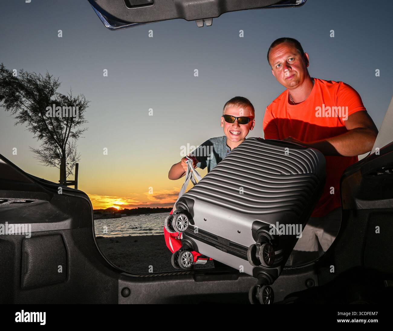Padre e figlio stanno caricando i bagagli nel bagagliaio della loro auto, pronti per una vacanza in famiglia su una splendida spiaggia durante il tramonto. Foto Stock
