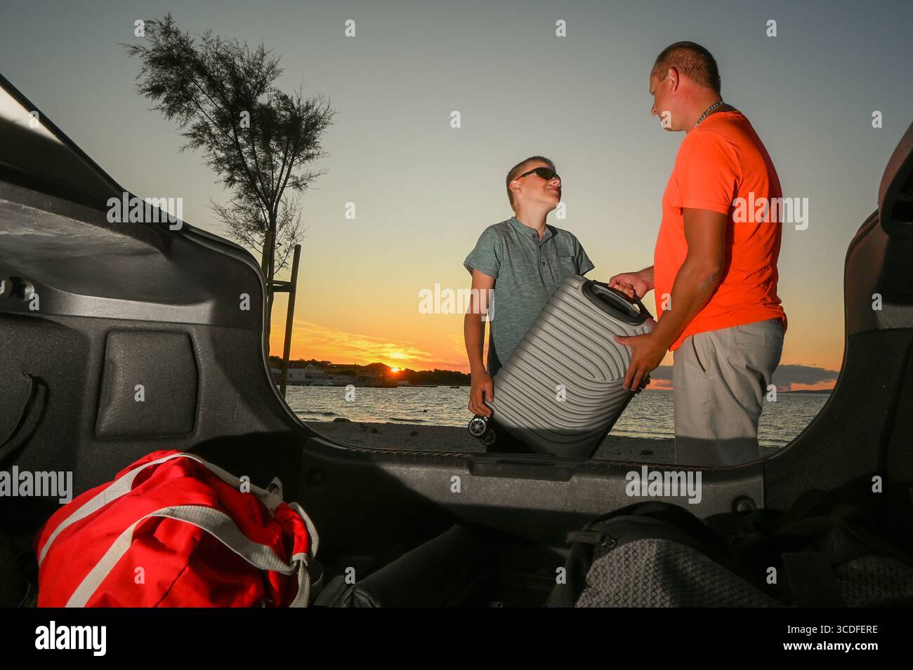 Padre e figlio stanno caricando i bagagli nel bagagliaio dell'auto, pronti per una vacanza al mare al tramonto. Foto Stock