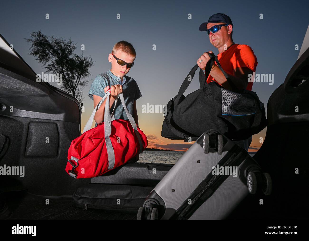 Padre e figlio stanno caricando i bagagli nel bagagliaio della loro auto, preparandosi per un viaggio in auto al tramonto. Foto Stock