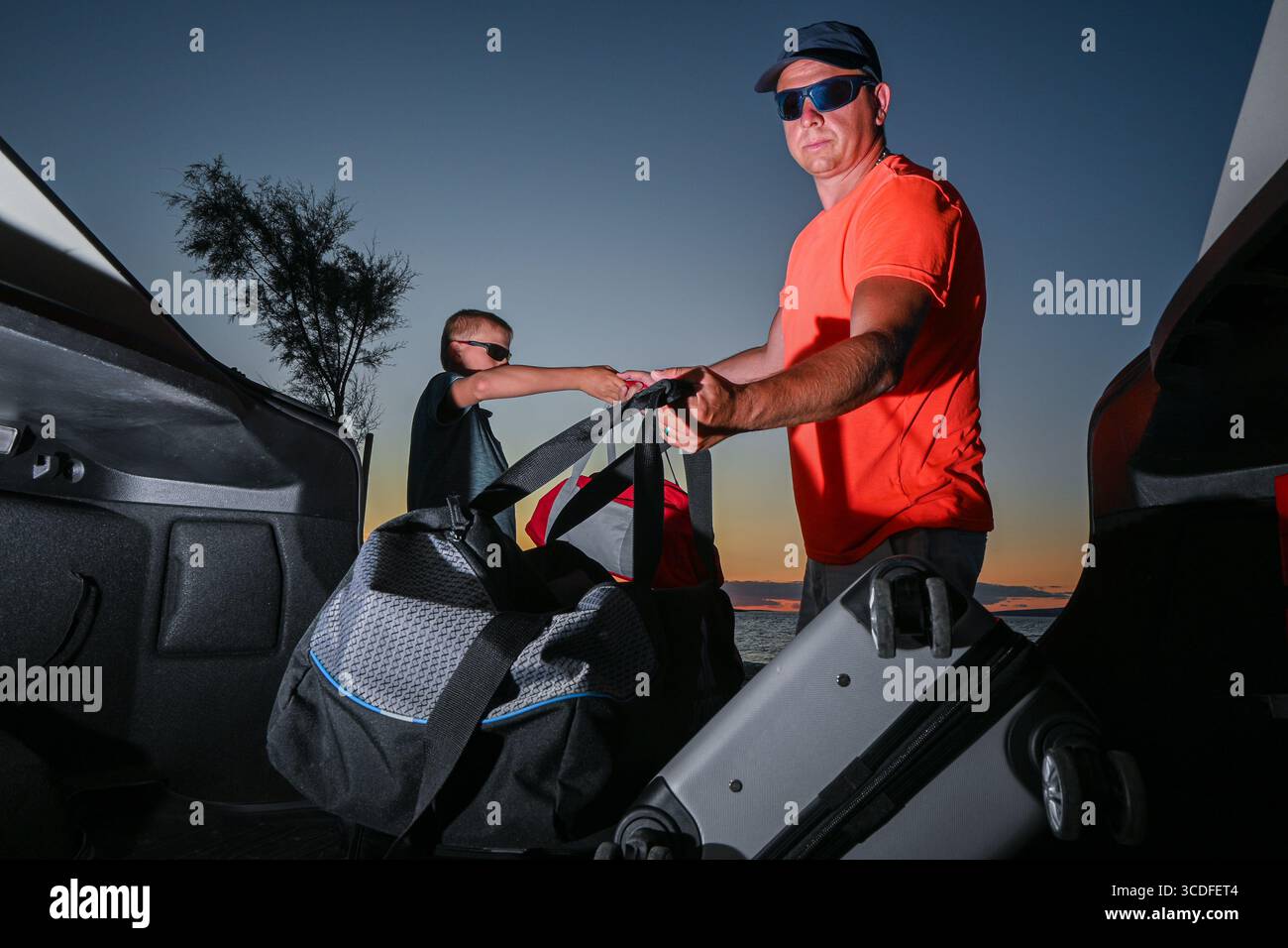 Padre e figlio stanno caricando i bagagli nel bagagliaio della loro auto, preparandosi per un viaggio in auto al tramonto. Foto Stock