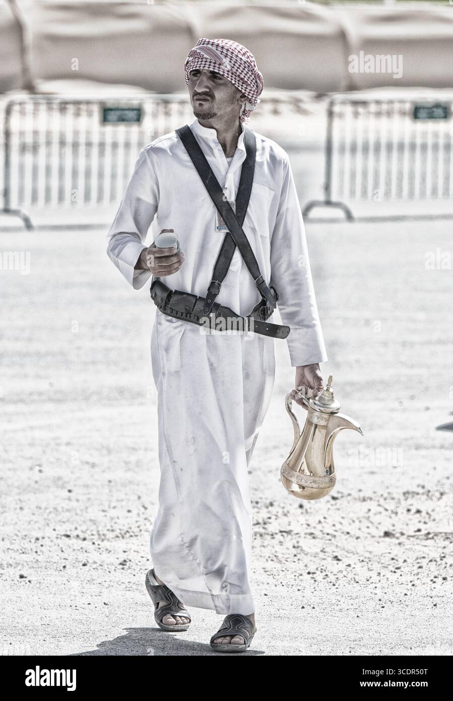 Uomo arabo in tradizionale tobe bianco e keffiyeh con caffettiera dallah dorata, patrimonio culturale, tradizione mediorientale, sfondo desertico. Foto Stock