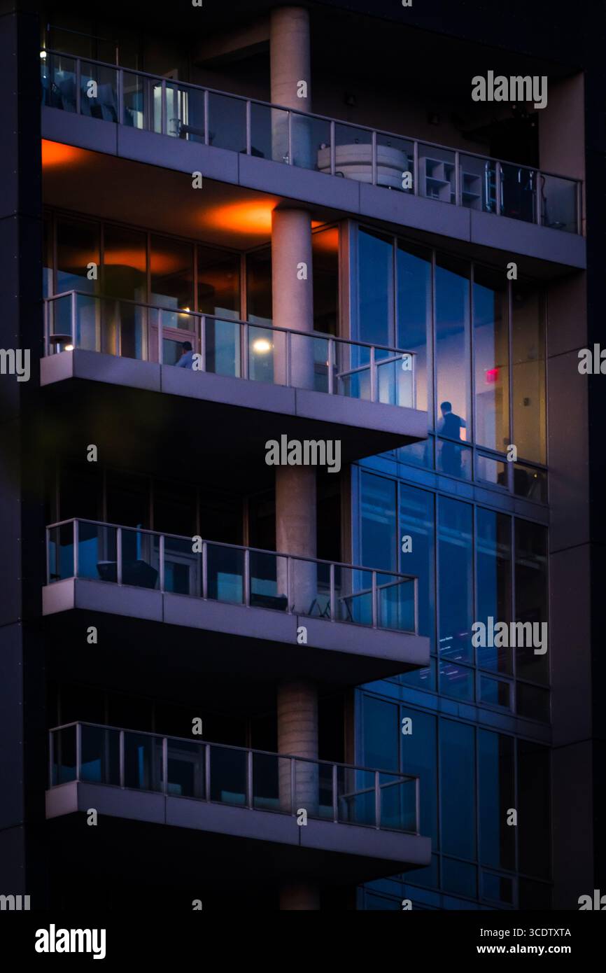 Vista di un alto edificio caratterizzato da balconi in vetro blu scuro e cemento, con calde luci arancioni che si illuminano dall'interno, creando un contrasto sorprendente, New York, New York, Stati Uniti. Foto Stock