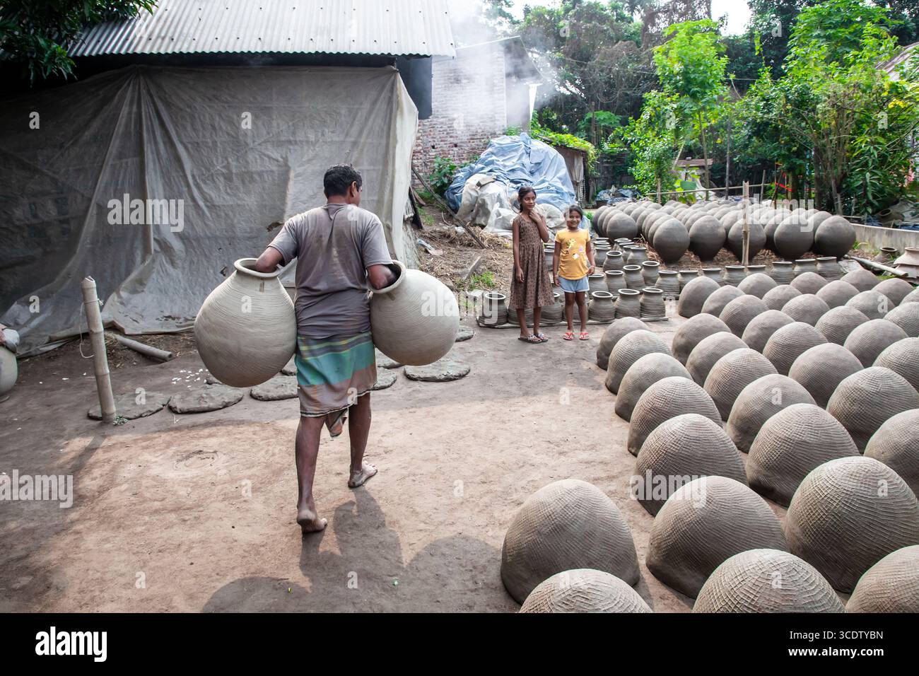 Munshiganj, Bangladesh - 23 novembre 2024: Vista di un vasaio che trasporta grandi vasi di argilla tra file di ceramica, con bambini che guardano nel giardino nebbioso e soleggiato. Foto Stock