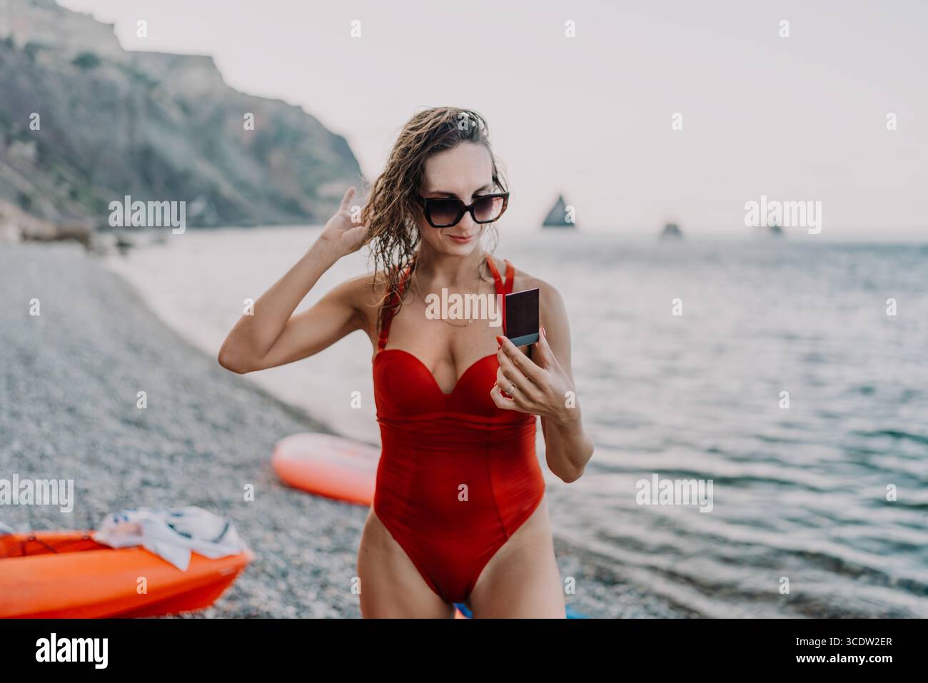Donna, spiaggia, foto istantanee. Giovane donna in costume da bagno rosso sulla spiaggia rocciosa che tiene una foto istantanea, regolando i capelli bagnati durante le vacanze estive. Foto Stock