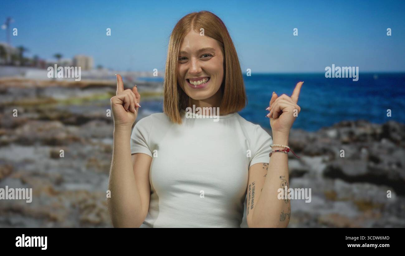 La giovane donna bionda punta le dita indice verso l'alto sulla costa rocciosa del mare soleggiato; ottimismo entusiasmo eccitazione gioia. Foto Stock