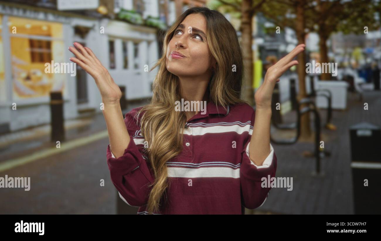 Donna alzata le mani per un gesto arrugginito in una strada pedonale dello shopping con una polo a righe; confusione dubbio frustrazione indecisione. Foto Stock