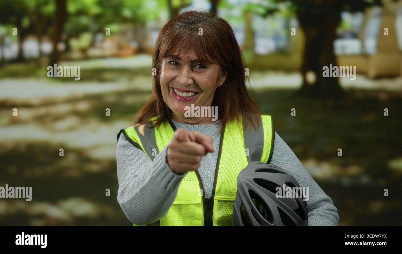 Una donna anziana ispanica sorride in un parco mentre punta la macchina fotografica, indossa un giubbotto riflettente e tiene in mano un casco da bicicletta, incarnando la sicurezza durante un Foto Stock