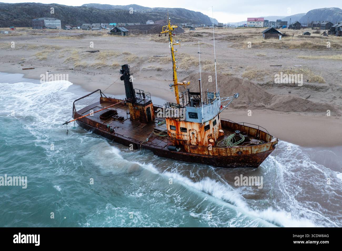 Vista aerea di un relitto arrugginito arenato su una spiaggia sabbiosa, onde che si infrangono dolcemente contro lo scafo intempestivo sullo sfondo di un remoto villaggio costiero, Teriberka, Oblast' di Murmansk, Russia. Foto Stock