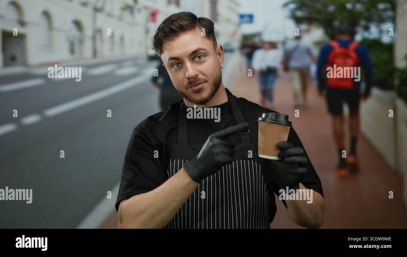 Il giovane con la barba nel grembiule punta alla tazza di caffè mentre si trova in strada, evidenziando il suo ruolo di cameriere con i guanti all'aperto in una città Foto Stock