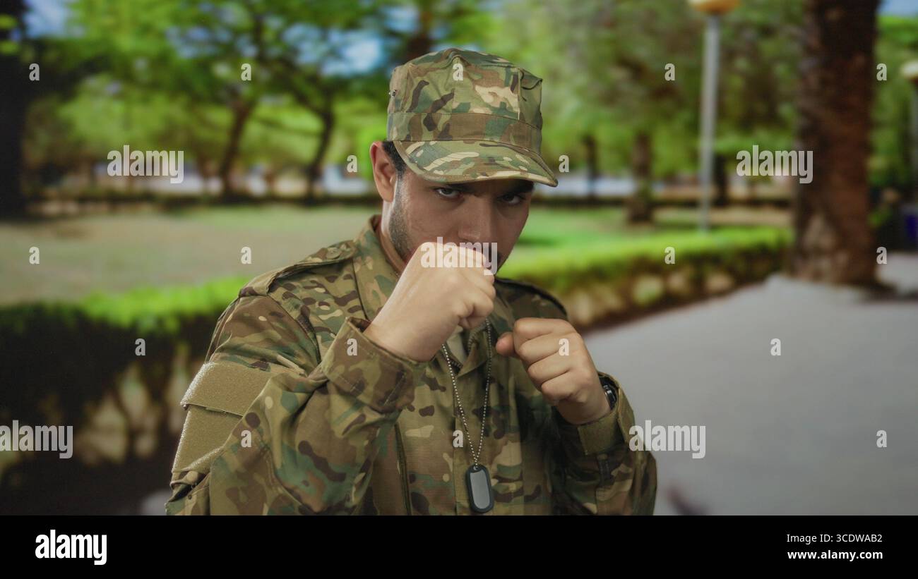 Uomo in uniforme mimetica solleva pugno armato in posa di combattimento in ambiente forestale con etichette per cani visibili; dovere coraggio. Foto Stock