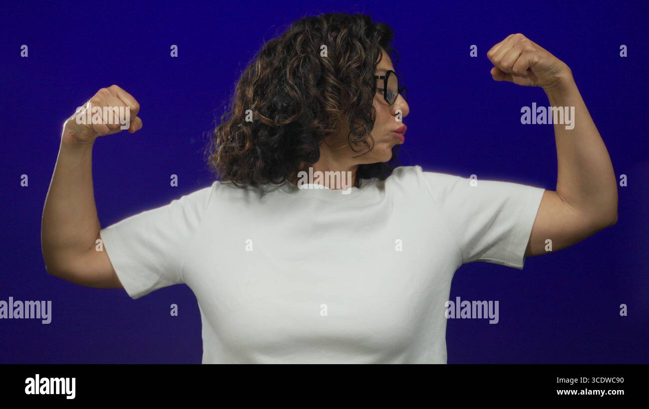 Donna ispanica di mezza età che flessa con sicurezza i muscoli contro un muro isolato di sfondo blu, mostrando forza e potenza nel suo casual Foto Stock