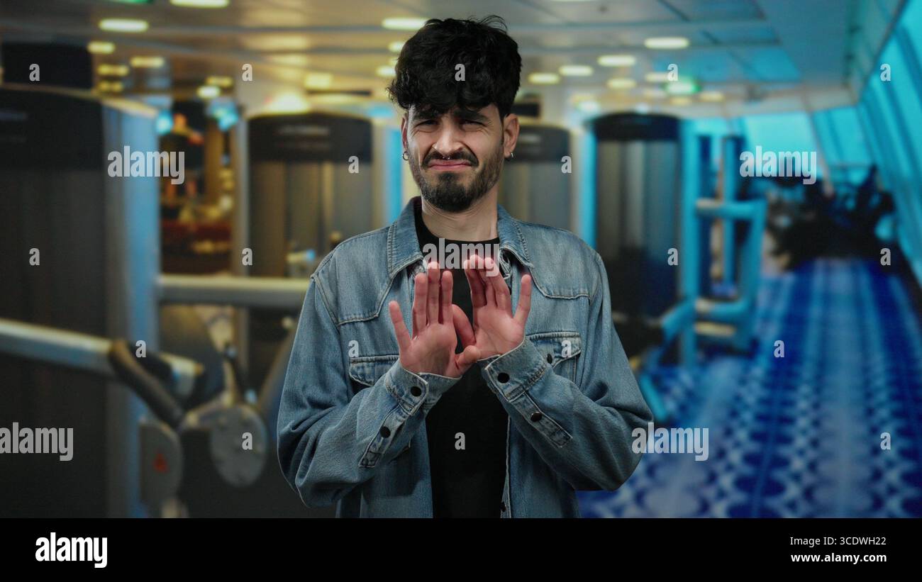 Giovane uomo che esprime disprezzo in palestra, indossa una giacca in denim sullo sfondo di moderne attrezzature fitness. Foto Stock