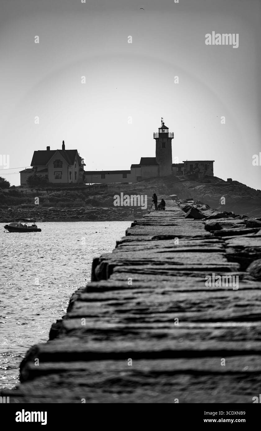 Una fotografia in bianco e nero cattura un molo roccioso che conduce verso una piccola isola con un faro ed edifici. Due figure che pescano sul molo Foto Stock