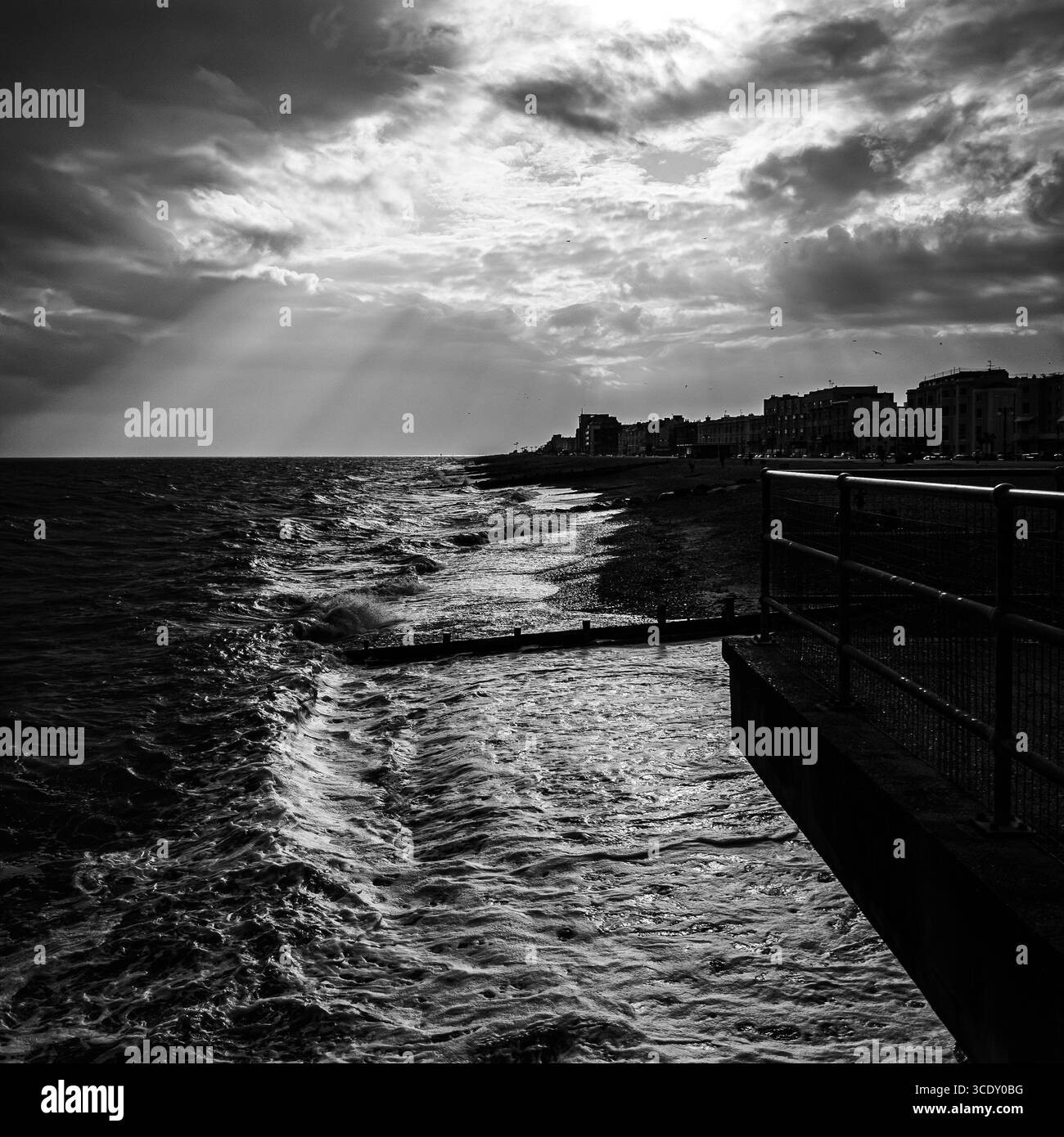 Un'immagine in bianco e nero dei raggi solari che illuminano il lungomare di worthing e i cavalli bianchi mentre le onde si infrangono sulla spiaggia - Immagine stock catturata con smartphone