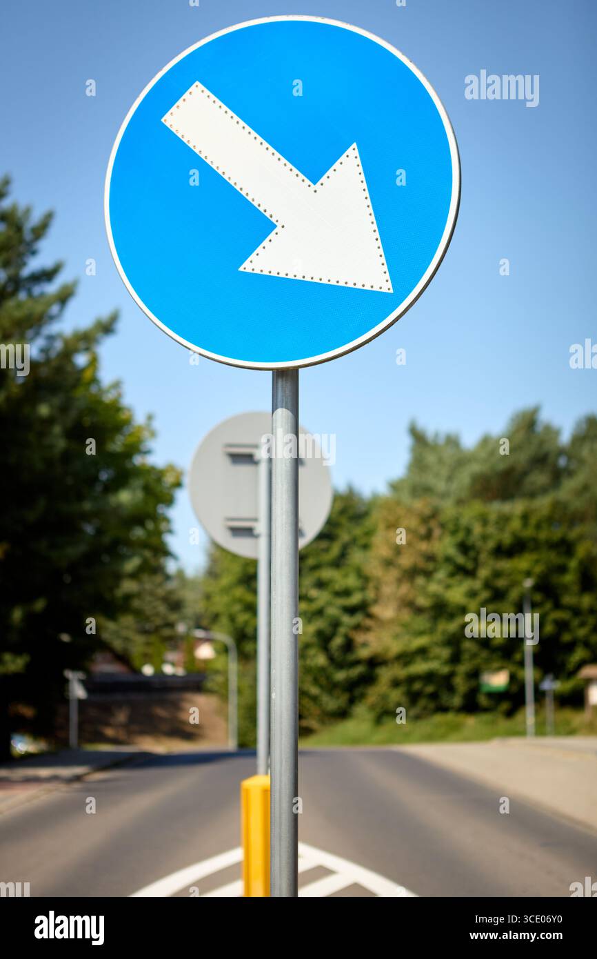 Circolare blu mantieni il cartello stradale a sinistra con una freccia bianca rivolta verso il basso a sinistra, posizionata accanto a una strada con alberi verdi sullo sfondo, i Foto Stock