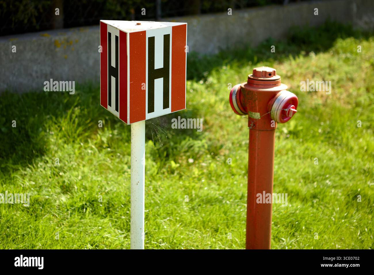 Idrante rosso vicino a un palo bianco con lati rossi e simbolo H nero, installato sull'erba verde vicino a un marciapiede di cemento, che indica la fonte d'acqua Foto Stock