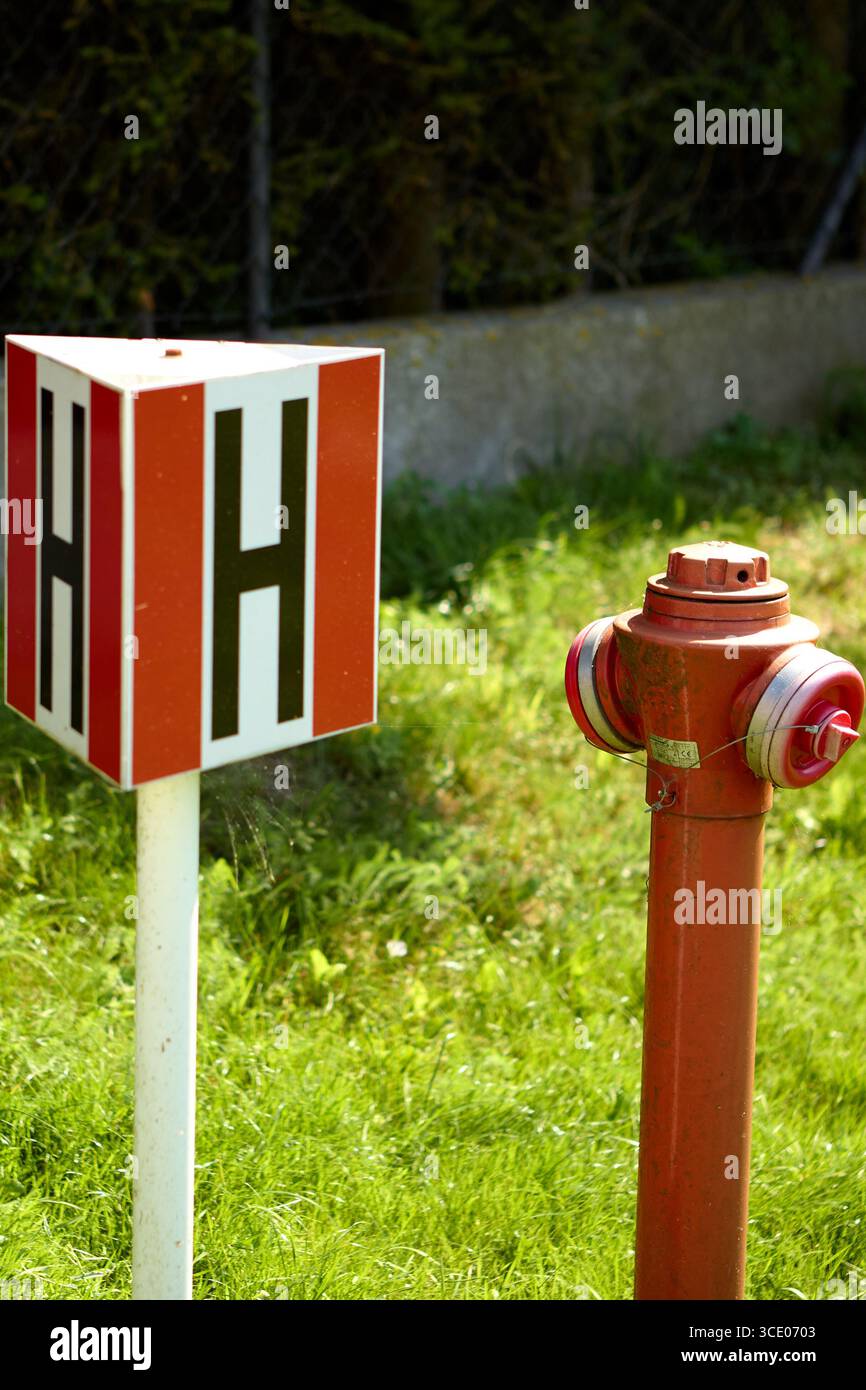 Idrante rosso vicino a un palo bianco con lati rossi e simbolo H nero, installato sull'erba verde vicino a un marciapiede di cemento, che indica la fonte d'acqua Foto Stock