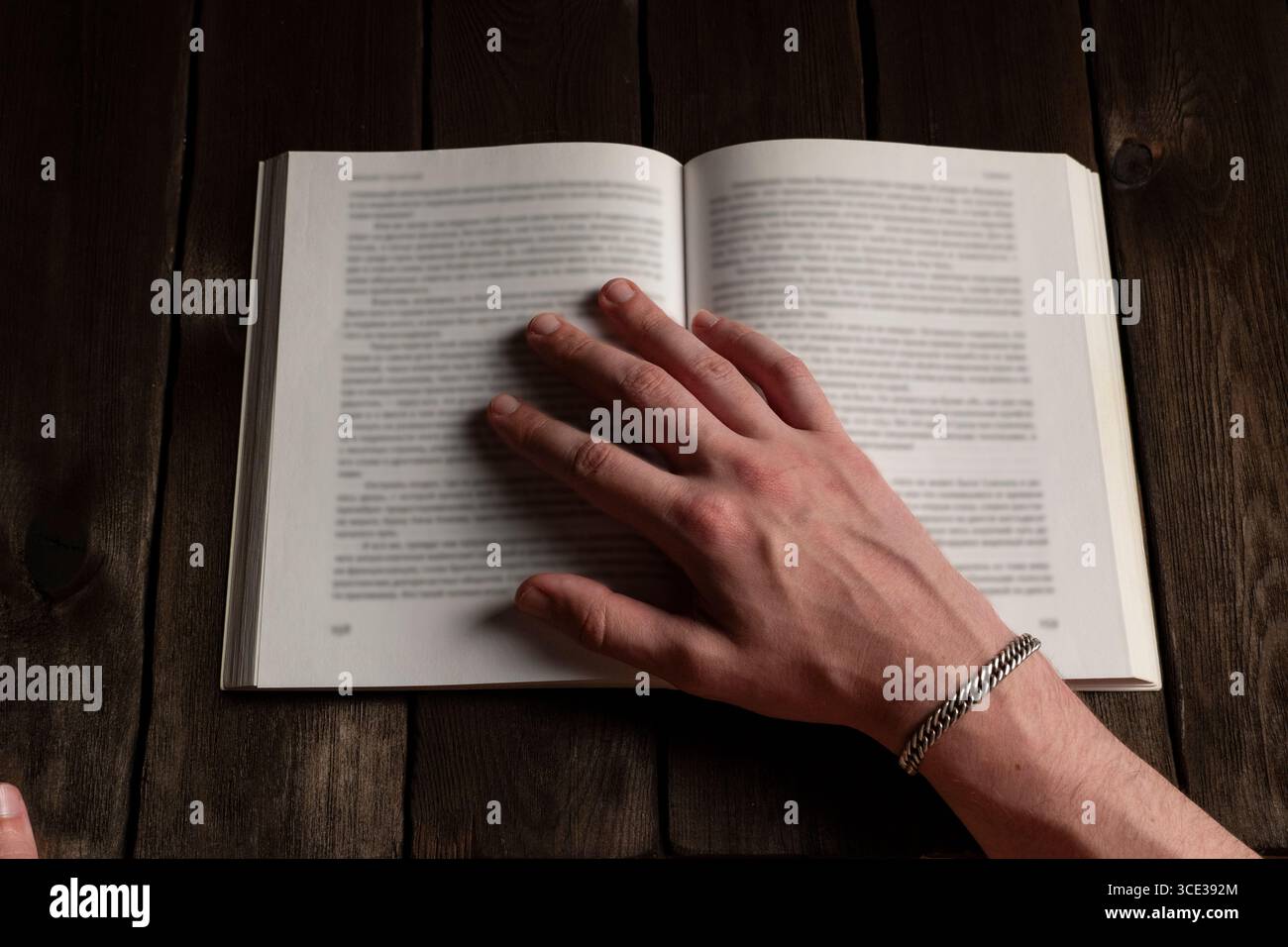 Un primo piano di una mano adornata da un braccialetto d'argento che poggia sulle pagine aperte di un libro su un rustico tavolo di legno, evocando un intimo bancomat di lettura e studio Foto Stock