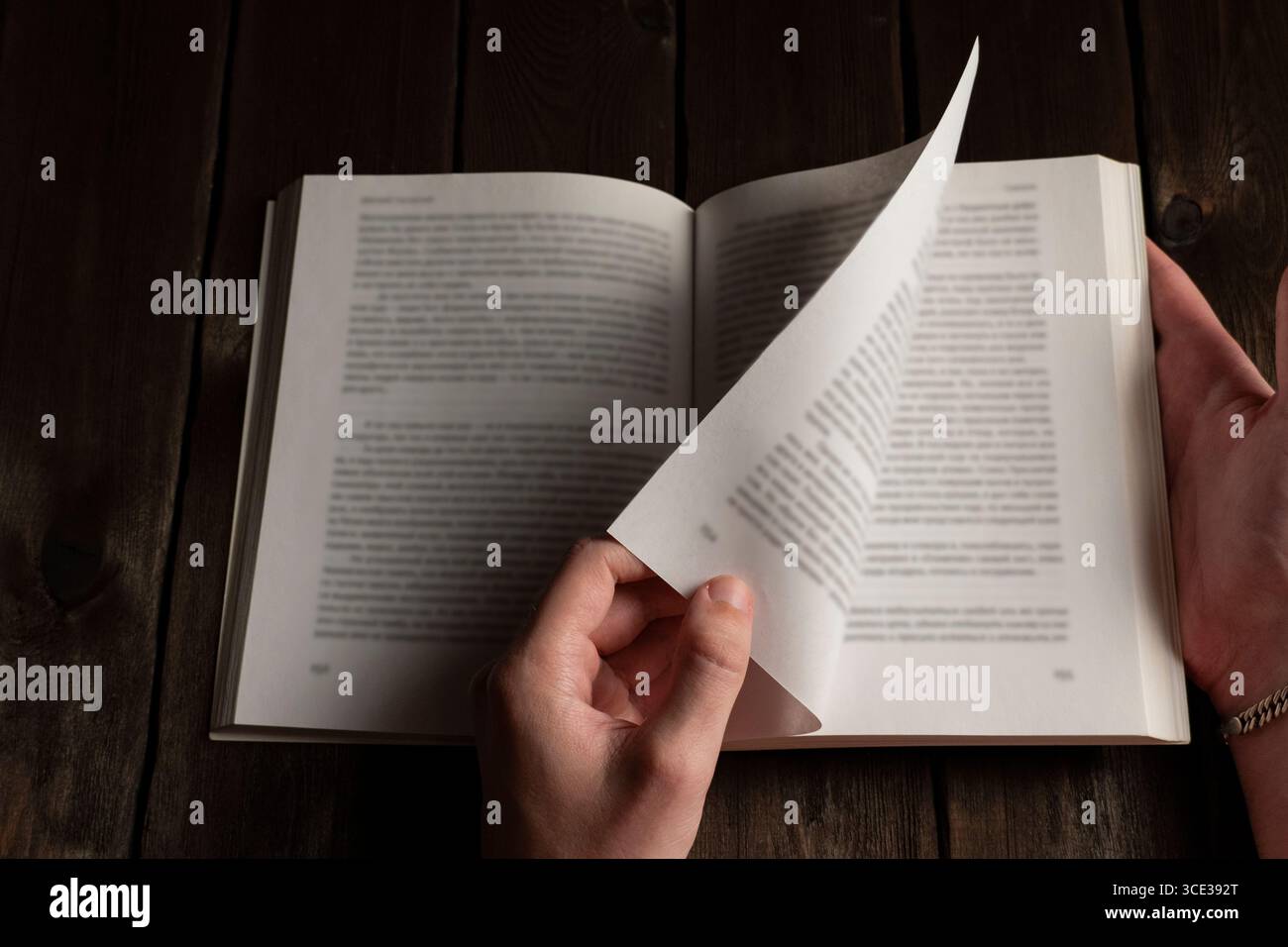 Le mani che sfogliano le pagine di un libro aperto su un tavolo di legno scuro, il testo sfocato evidenzia il movimento della lettura e l'esperienza tattile Foto Stock