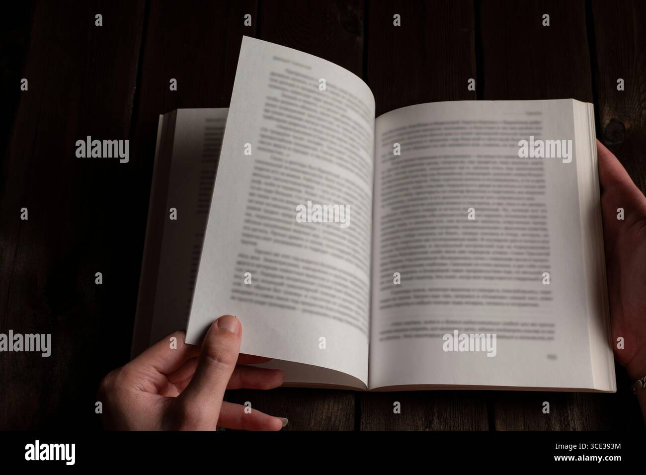 Una persona che gira una pagina di un libro aperto su un tavolo di legno scuro, con le mani a fuoco, catturando un'atmosfera accogliente e studiosa perfetta per leggere e leggere Foto Stock