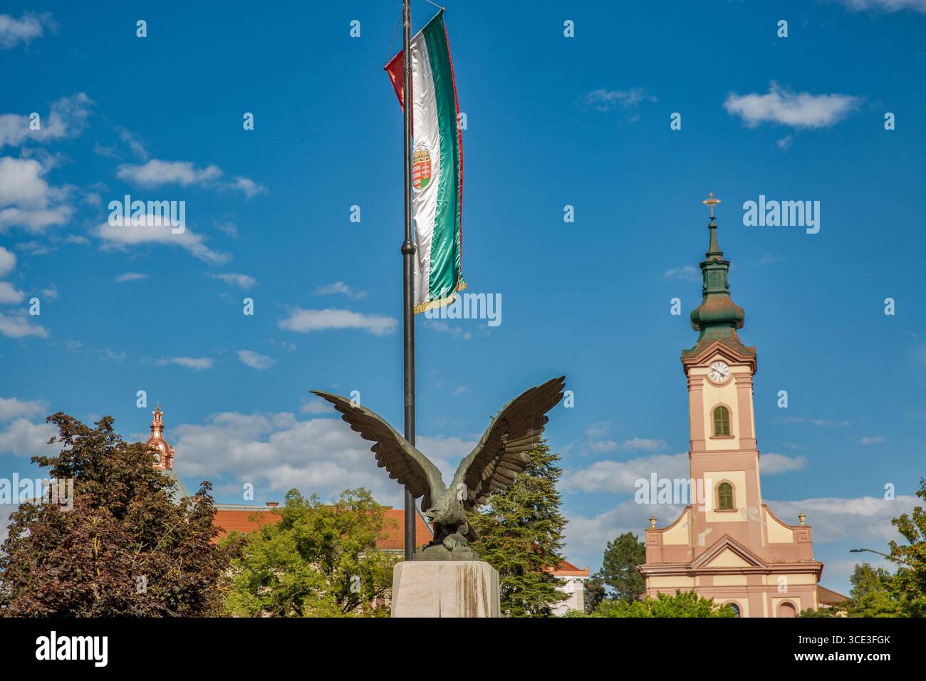 Statua in bronzo dell'aquila di Turul con ali sparse, posizionata sotto un'asta con la bandiera ungherese, adagiata su un cielo blu brillante e un monumento storico Foto Stock