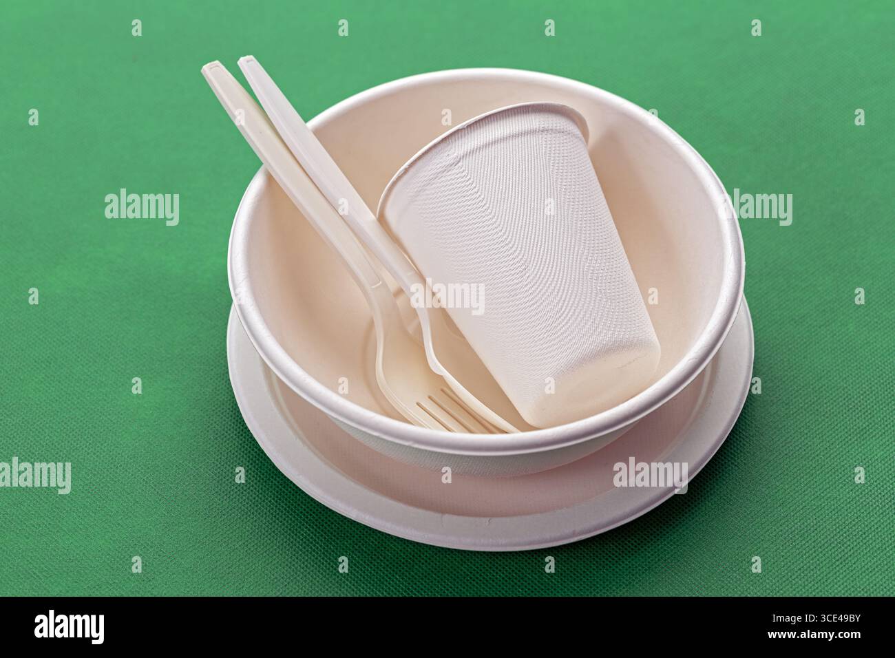 Posate biodegradabili, tazze e piatti su sfondo verde, che offrono opzioni gastronomiche ecologiche. utensili e piatti bianchi sottolineano Foto Stock