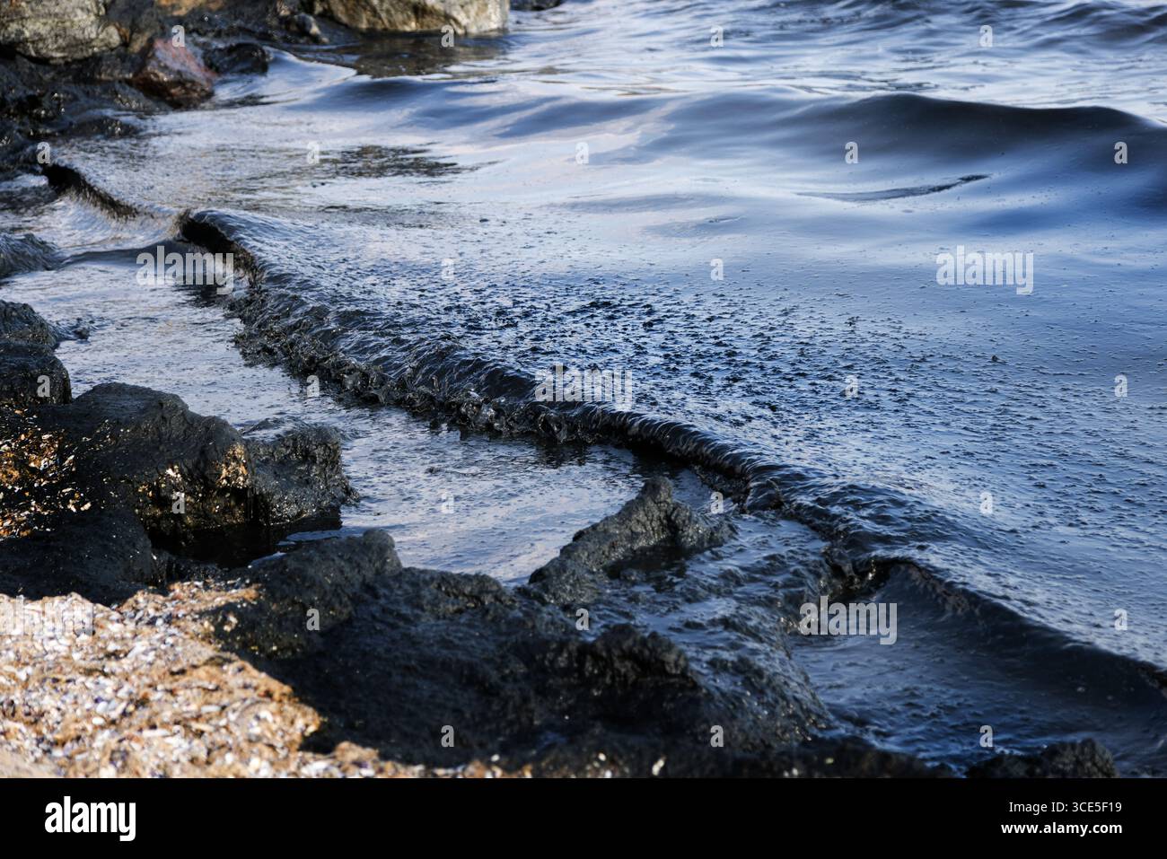 Costa rocciosa colpita da onde scure ricoperte di arcobaleno di fuoriuscite di petrolio, simbolo di disastro ambientale e minaccia per la vita marina causata dall'acqua Foto Stock