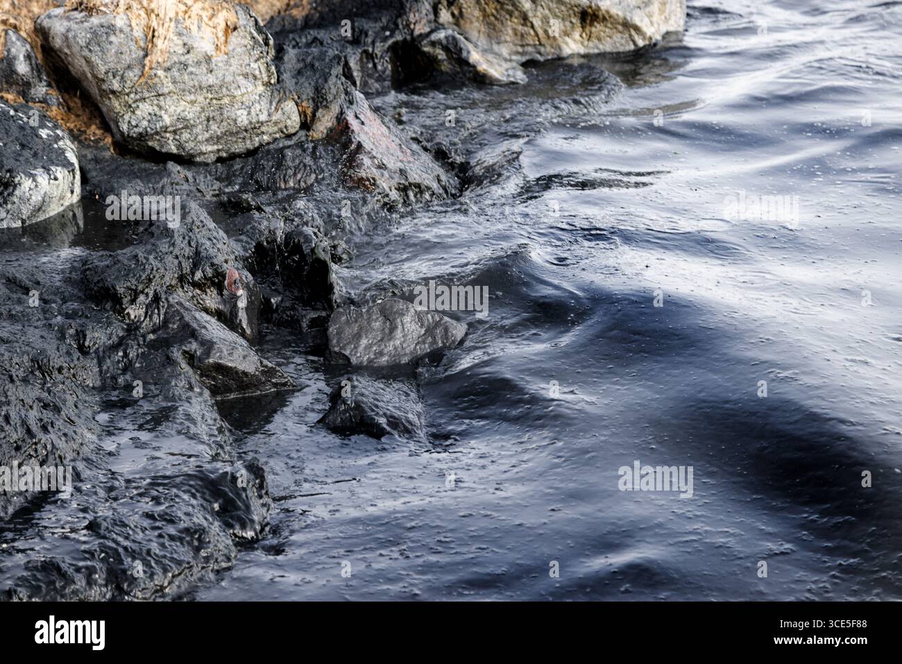 Costa rocciosa colpita da onde scure ricoperte di arcobaleno di fuoriuscite di petrolio, simbolo di disastro ambientale e minaccia per la vita marina causata dall'acqua Foto Stock