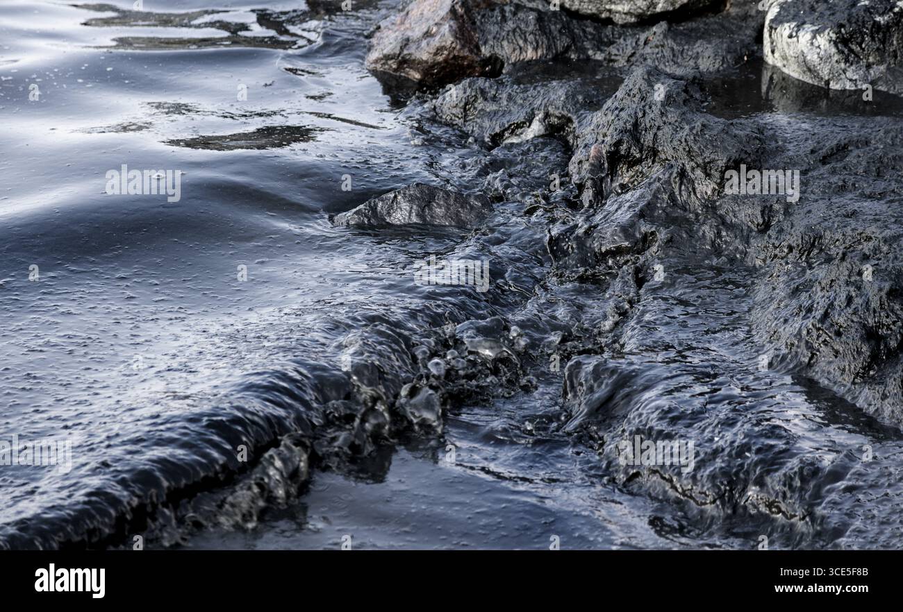 Costa rocciosa colpita da onde scure ricoperte di arcobaleno di fuoriuscite di petrolio, simbolo di disastro ambientale e minaccia per la vita marina causata dall'acqua Foto Stock