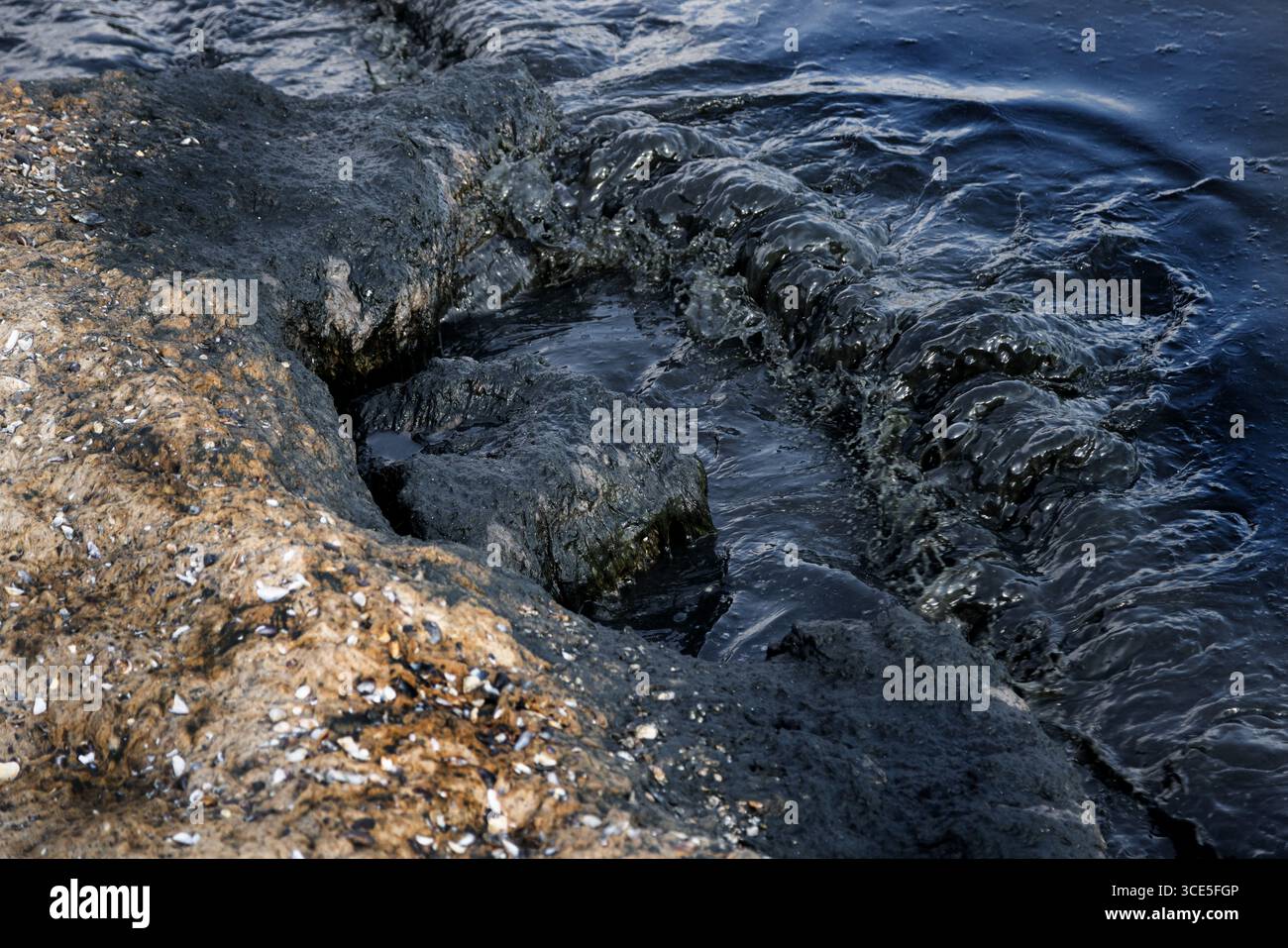 Costa rocciosa colpita da onde scure ricoperte di arcobaleno di fuoriuscite di petrolio, simbolo di disastro ambientale e minaccia per la vita marina causata dall'acqua Foto Stock