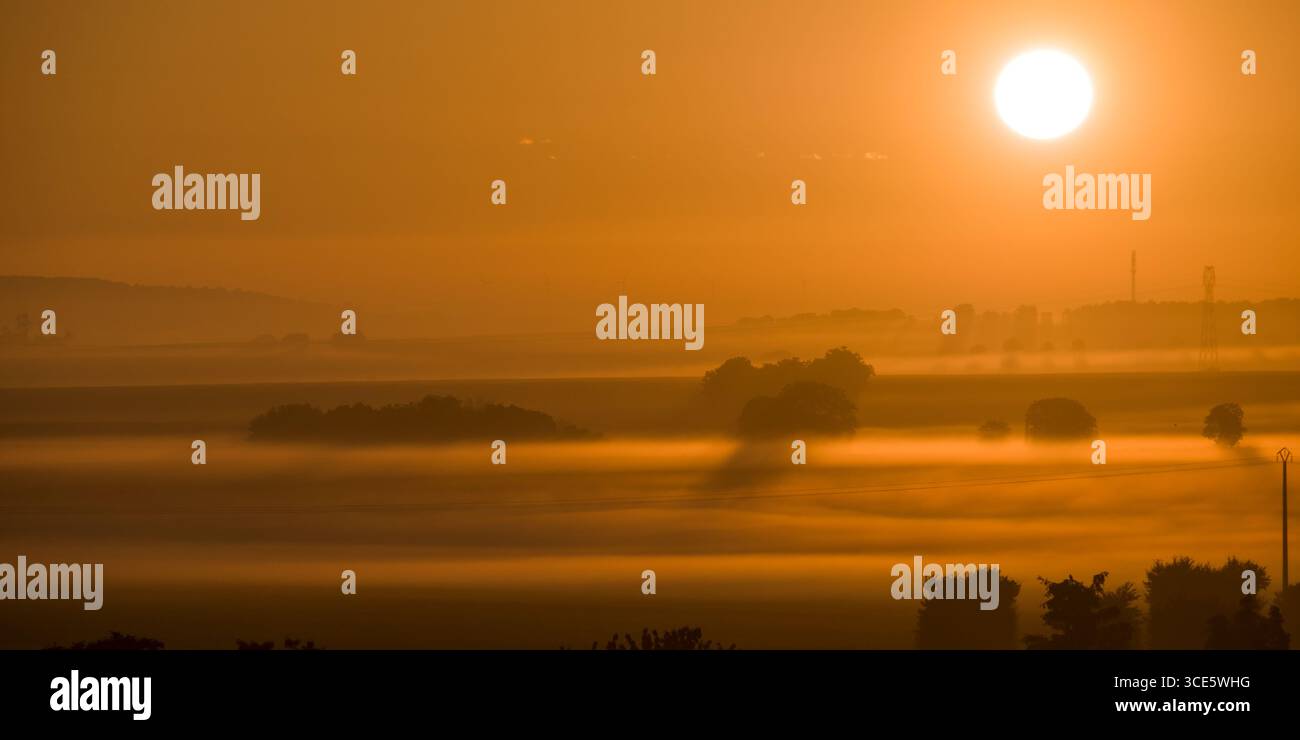 Alba sulla campagna francese con campi ondulati, alberi sparsi e nebbia mattutina, inondati dalla calda luce arancione Foto Stock