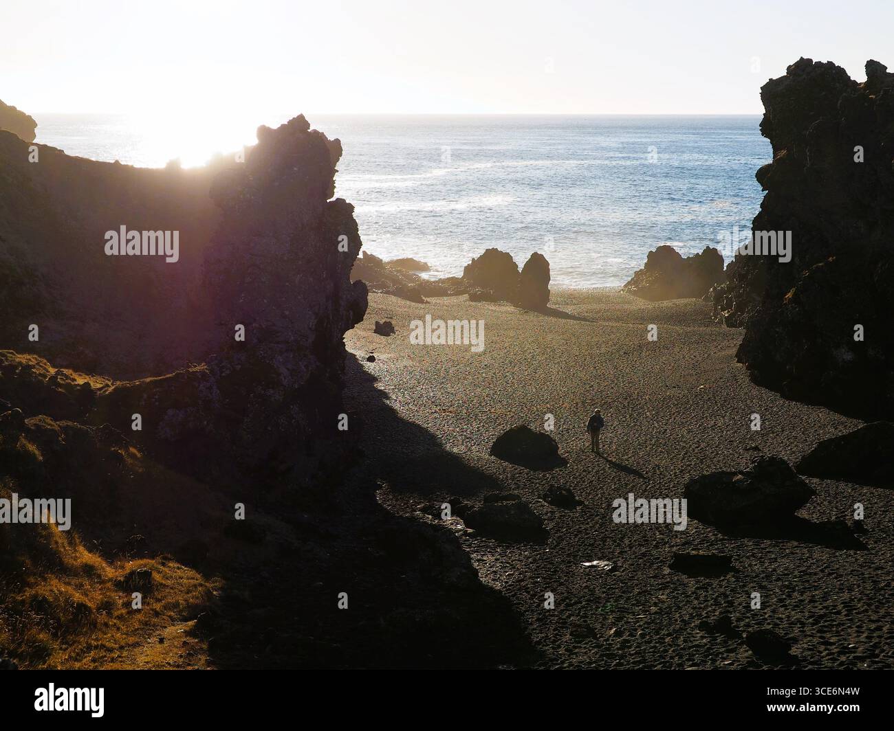 Uomo che cammina su una spiaggia di sabbia nera sotto i raggi del sole, con una spettacolare sagoma di rocce e ombre: Avventura, sfida, opportunità, esplorazione del futuro Foto Stock