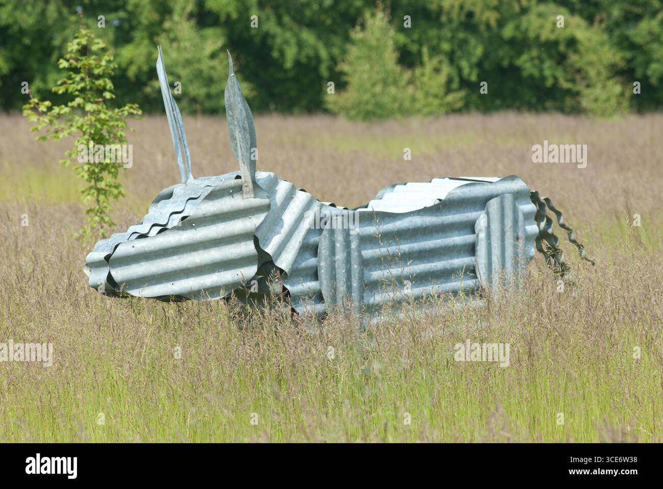 Una scultura di mucca metallizzata riposa tranquillamente in un alto campo erboso, circondato dalla natura in una giornata di sole, che mostra l'arte rurale. Foto Stock
