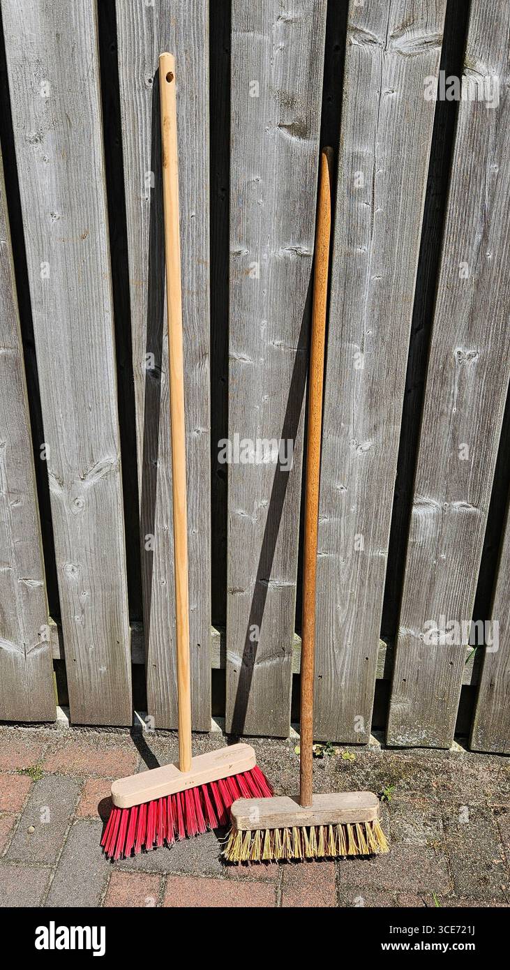 Due scope con manici in legno e setole colorate appoggiate su una recinzione in legno rustica su un patio pavimentato, per catturare un momento semplice. - Immagine stock catturata con smartphone