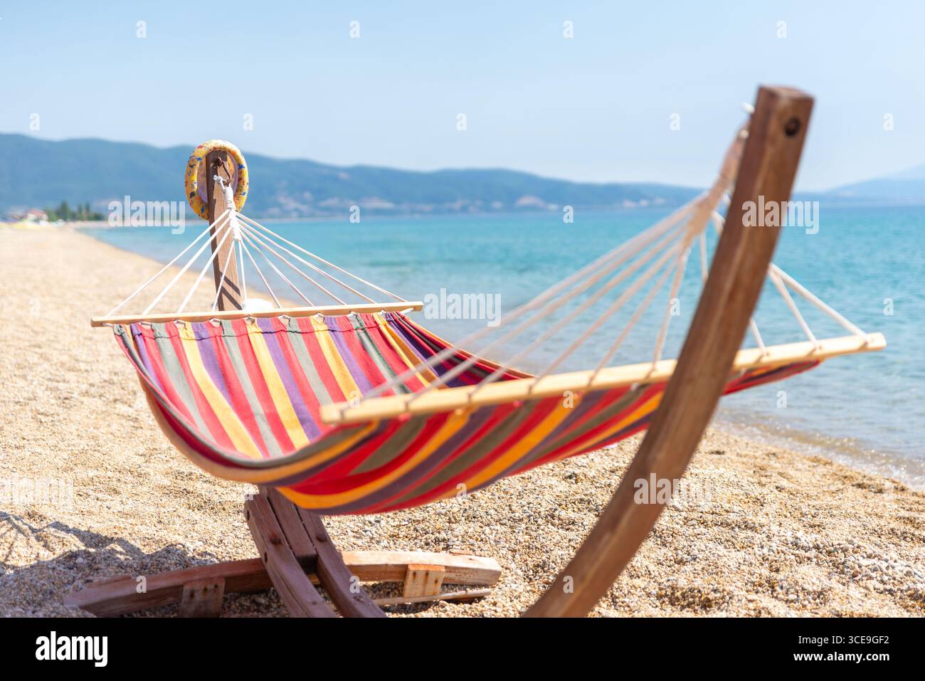 Vista completa di un'amaca colorata a righe su una tranquilla spiaggia di ciottoli sul mare, simbolo del tempo libero estivo Foto Stock