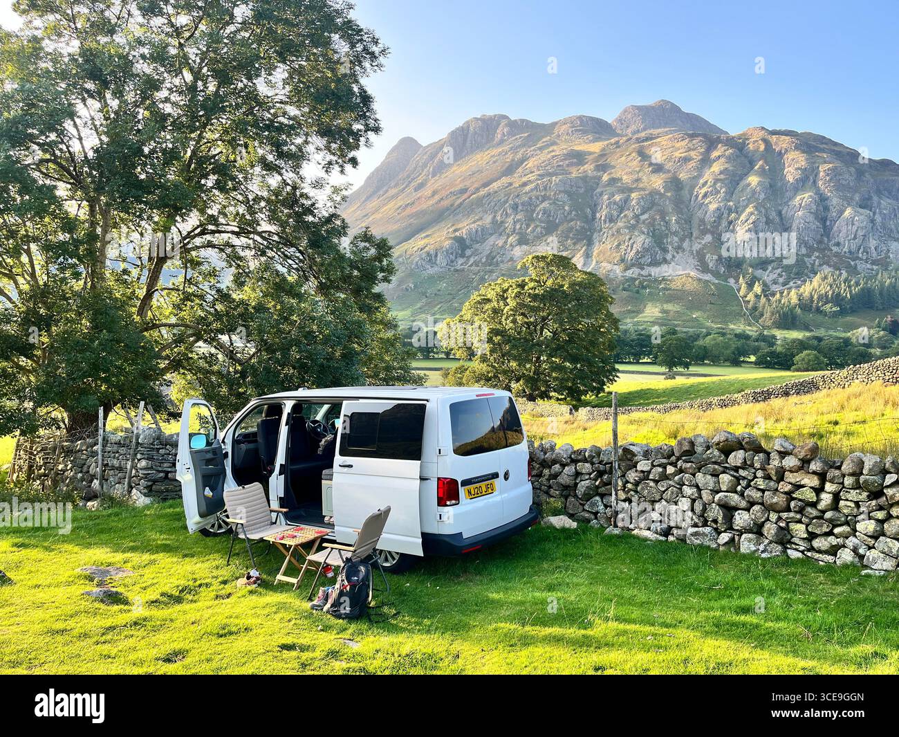 Un camper bianco VW Transporter parcheggiato sul lato della strada nel Lake District con montagne sullo sfondo e una sedia e un tavolo all'esterno. - Immagine stock catturata con smartphone