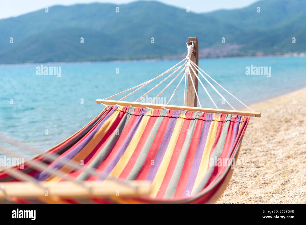 Amaca su una spiaggia di ciottoli con acque turchesi e montagne verdi lontane, perfetta per il relax Foto Stock
