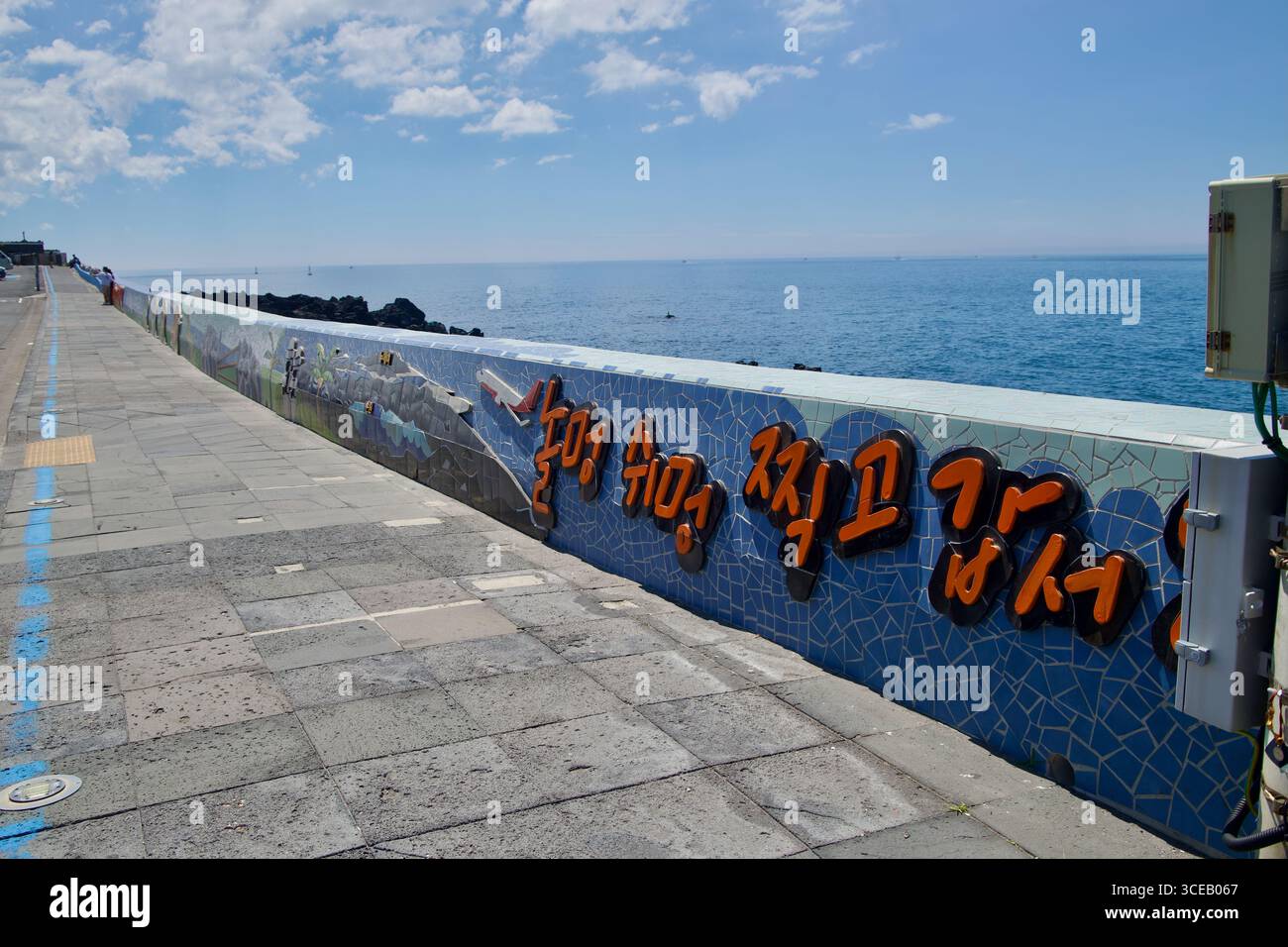 Una diga a mosaico piastrellato con scritta coreana arancione rialzata confina con la passeggiata a Eoyeong Park nella città di Jeju, affacciata sulle rocce basaltiche e sulle aperte Foto Stock
