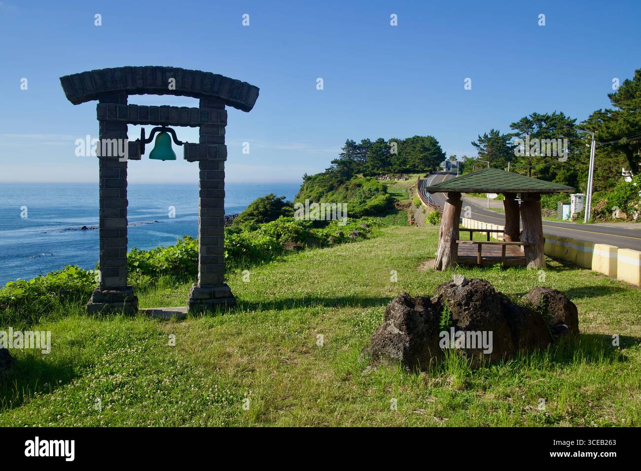 Un piccolo parco sulla cima della scogliera sulla strada costiera di Hagu-Aewol presenta una campana incastonata in un arco di basalto e un rustico rifugio in legno che si affaccia sul mare aperto e sul vin Foto Stock