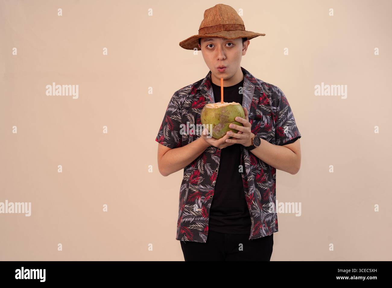 uomo in camicia stampata che regge noce di cocco con paglia arancione e indossa un cappello di paglia Foto Stock