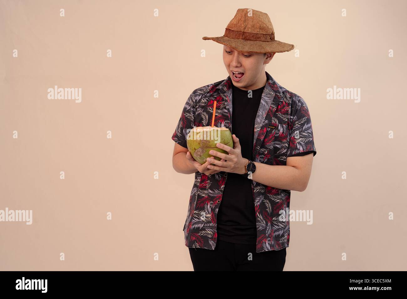 uomo in camicia stampata che regge noce di cocco con paglia arancione e indossa un cappello di paglia Foto Stock