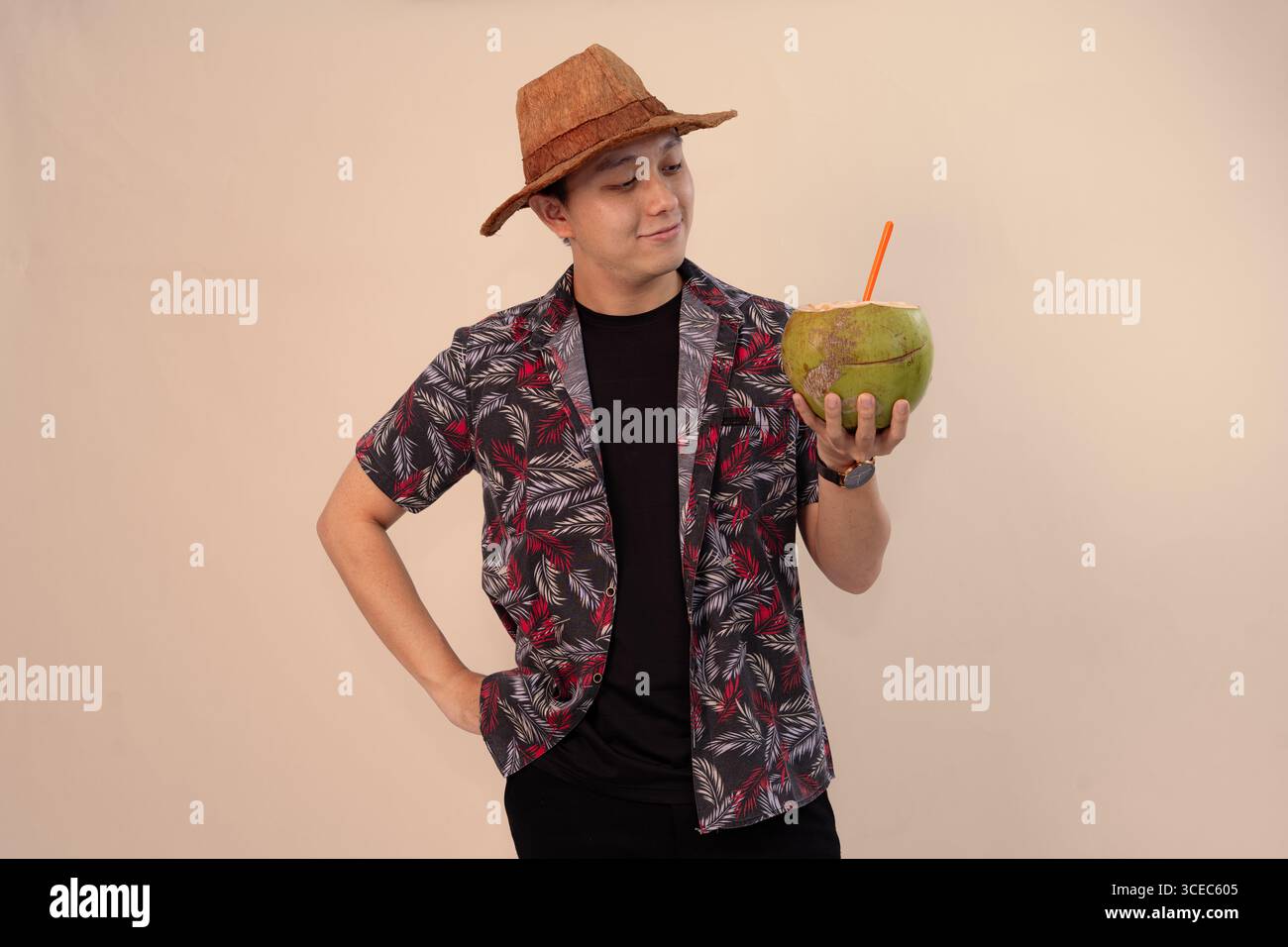 uomo in camicia stampata che regge noce di cocco con paglia arancione e indossa un cappello di paglia Foto Stock
