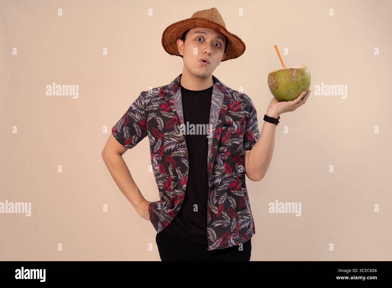 uomo in camicia stampata che regge noce di cocco con paglia arancione e indossa un cappello di paglia Foto Stock