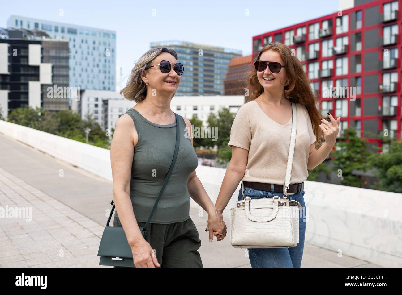Madre e figlia camminano mano nella mano all'aperto, sorridono e indossano occhiali da sole. L'ambiente urbano e' vivace, sfoggia uno stile di vita moderno e. Foto Stock