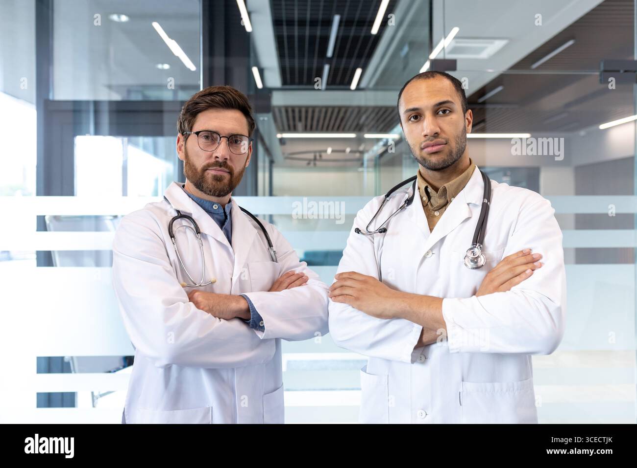 Due professionisti medici maschi in piedi con le braccia incrociate, indossando cappotti bianchi e stetoscopi, trasudando fiducia e professionalità in un ufficio moderno. Questa immagine simboleggia l'assistenza sanitaria, il lavoro di squadra e la fiducia. Foto Stock