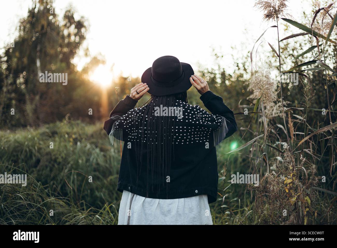 Una donna sta in un campo, rivolto verso l'esterno, tenendo il cappello mentre il sole tramonta. Il caldo bagliore del tramonto mette in risalto la sua silhouette, catturando una mamma serena Foto Stock