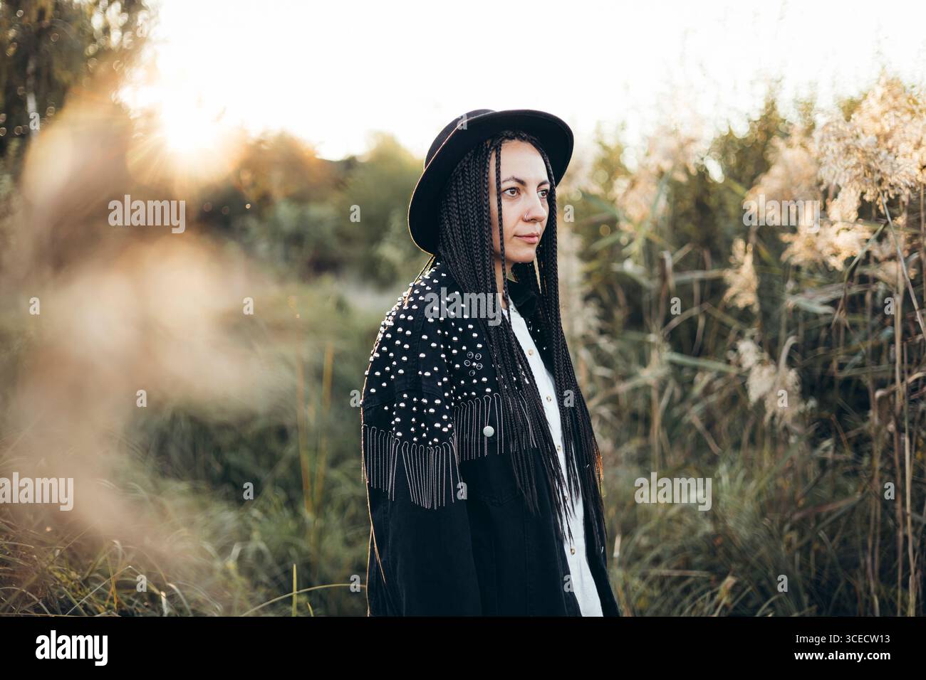 Una donna si trova in un campo tranquillo al tramonto, indossando un elegante cappello e capelli intrecciati. Il caldo bagliore del sole crea un'atmosfera tranquilla e contemplativa Foto Stock