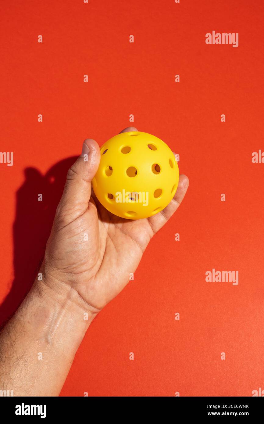 La mano corta e irriconoscibile regge un picchetto giallo vibrante su un audace sfondo arancione in uno studio. La figura anonima enfatizza il playfu Foto Stock