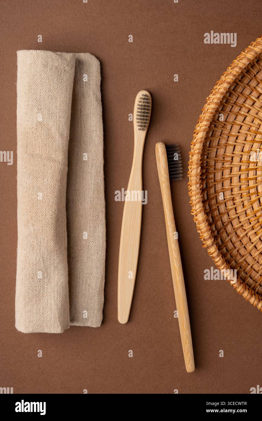 Vista dall'alto di due spazzolini da denti in bambù insieme a un asciugamano di lino beige e un cestello in vimini, su sfondo marrone in tessuto, per un ambiente ecologico Foto Stock